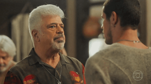A Man With White Hair And A Beard Stands Next To Another Man With A Necklace Around His Neck GIF