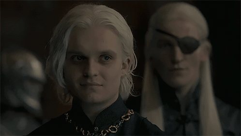 A Man With White Hair And A Necklace Is Standing Next To Another Man With A Blindfold On His Eye GIF