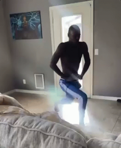 A Man Without A Shirt Is Dancing In A Living Room With A Couch . GIF