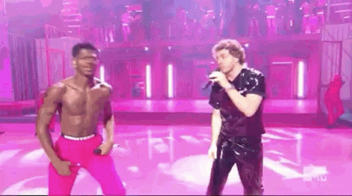 A Man Without A Shirt Is Singing Into A Microphone While Another Man Stands Next To Him On A Stage . GIF