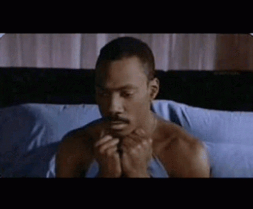 a man without a shirt is sitting on a bed with his hands folded in prayer .