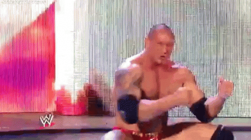 A Man Without A Shirt Is Sitting On A Stage In Front Of A Large Screen With The Letter W On It . GIF