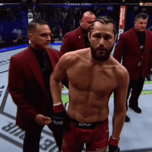 A Man Without A Shirt Is Standing In A Boxing Ring Surrounded By Men In Red Suits . GIF