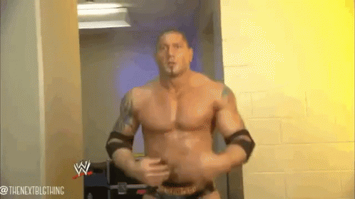 A Man Without A Shirt Is Standing In A Hallway With A Wrestling Logo On The Wall Behind Him . GIF