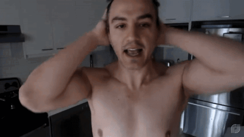 a man without a shirt is standing in a kitchen with his hands on his head .