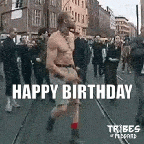A Man Without A Shirt Is Walking Down A Street In Front Of A Crowd Of People . GIF