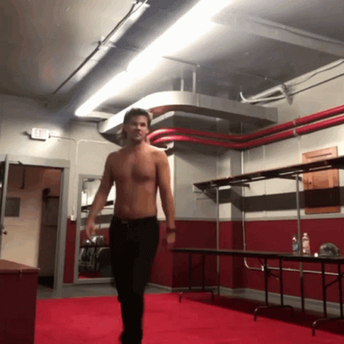 A Man Without A Shirt Is Walking In A Room With An Exit Sign On The Wall GIF