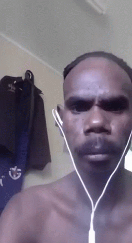 A Man Without A Shirt Is Wearing Ear Buds In Front Of A Wall With Clothes Hanging GIF