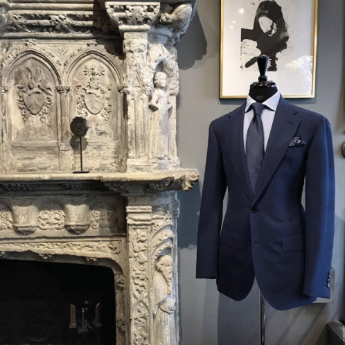 A Mannequin Wearing A Blue Suit And Tie Stands In Front Of A Stone Fireplace GIF