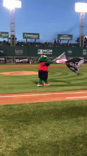 A Mascot Holding A Flag On A Baseball Field With A Mgm Sign Behind Him GIF