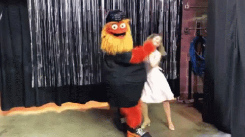 A Mascot Is Dancing With A Woman In A White Dress In Front Of A Black Curtain GIF