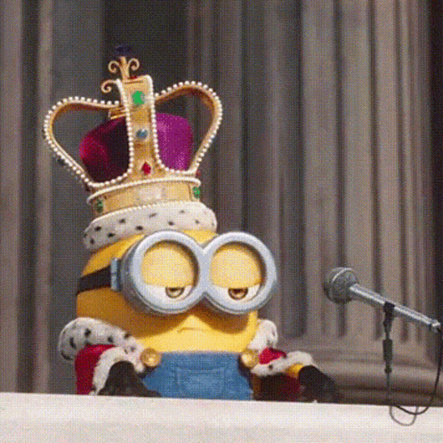 A Minion Wearing A Crown And Goggles Is Sitting At A Podium In Front Of A Microphone GIF