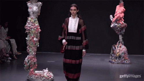 A Model Walks Down The Runway At A Fashion Show In Front Of Sculptures GIF
