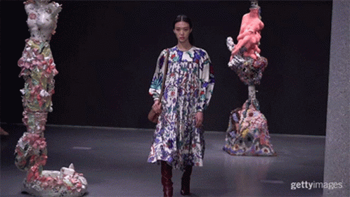 A Model Walks Down The Runway At A Fashion Show In Front Of Statues GIF