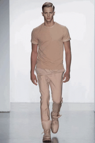 A Model Walks Down The Runway At A Fashion Show Wearing A Tan T-Shirt And Tan Pants GIF