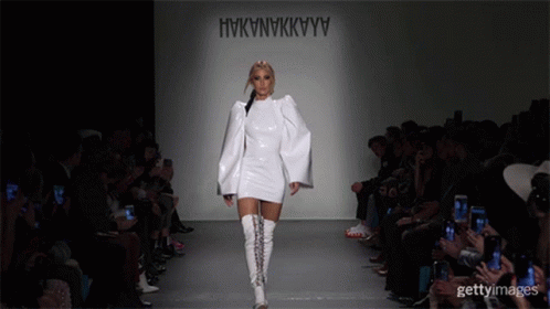 A Model Walks Down The Runway At A Fashion Show Wearing A White Dress And White Boots GIF