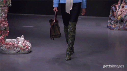 A Model Walks Down The Runway At A Fashion Show While Holding A Purse And Wearing Green Boots GIF