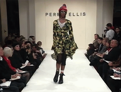 A Model Walks Down The Runway In Front Of A Wall That Says Perri Ellis GIF