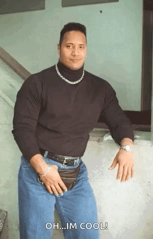 A Muscular Man Wearing A Black Turtleneck And Jeans Is Standing In Front Of A Wall . GIF