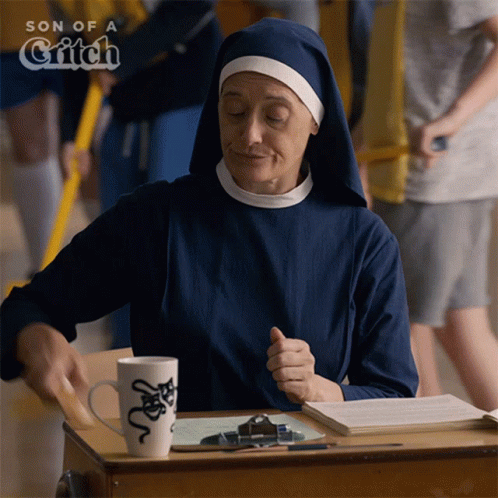 A Nun Sits At A Desk In Front Of A Sign That Says Son Of A Witch GIF