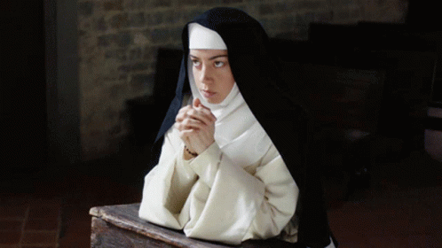 A Nun Sits On A Wooden Table With Her Hands Folded GIF