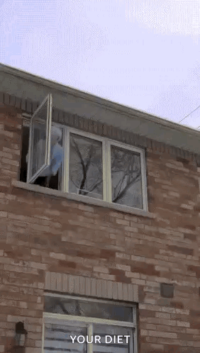 A Person In A Blue Hoodie Is Jumping Out Of A Window Of A Brick House . GIF