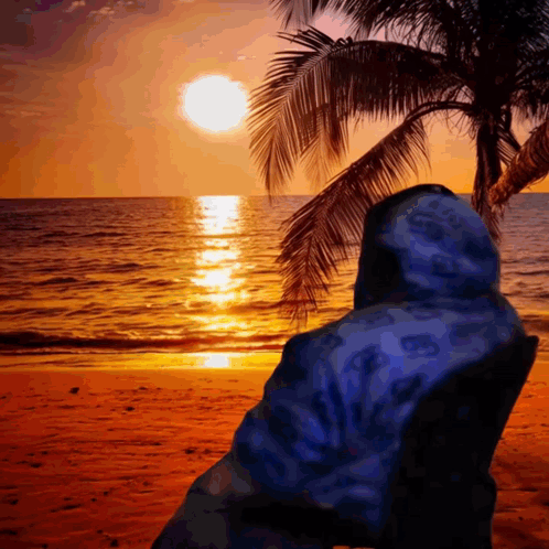 A Person In A Blue Hoodie Sits On A Beach Watching The Sun Set GIF