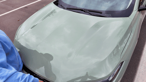 A Person In A Blue Sweatshirt Is Looking Under The Hood Of A Green Car GIF