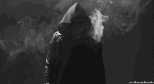 A Person In A Hooded Jacket Is Smoking A Cigarette In A Dark Room GIF