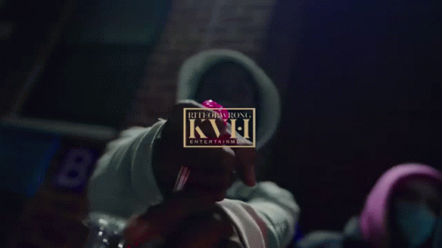 A Person In A Hoodie Holds A Red Object In Front Of A Sign That Says Kvh GIF