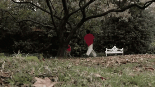 A Person In A Red Shirt Is Running In A Park Near A White Bench GIF