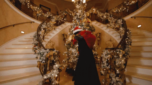 A Person In A Santa Hat Is Standing On A Staircase With Christmas Decorations GIF