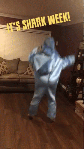 A Person In A Shark Costume Is Dancing In A Living Room With A Sign That Says It 'S Shark Week GIF