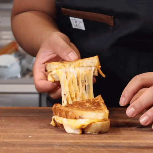 A Person In An Apron Is Holding A Grilled Cheese Sandwich With Cheese Coming Out Of It GIF