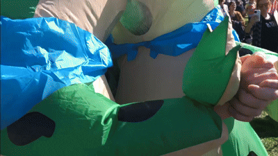 A Person In An Inflatable Costume With A Blue Cape Holds Another Person 'S Hand GIF