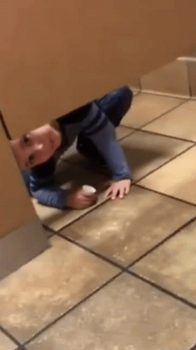 A Person Is Crawling Under A Wall In A Public Restroom Holding A Cup . GIF