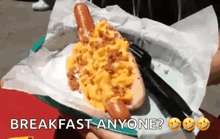 A Person Is Holding A Hot Dog With Scrambled Eggs On It And The Words Breakfast Anyone Below It GIF