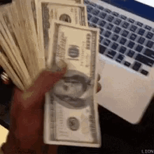 A Person Is Holding A Large Stack Of Money In Front Of A Laptop . GIF