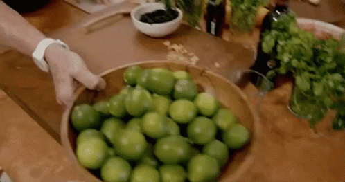 A Person Is Holding A Wooden Bowl Filled With Limes On A Table . GIF