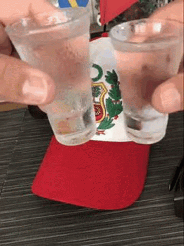 A Person Is Holding Two Shot Glasses In Front Of A Flag . GIF