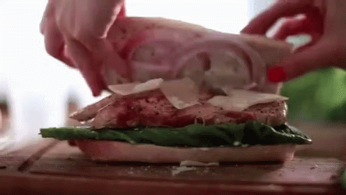a person is making a sandwich with chicken , lettuce , cheese and onions on a cutting board .