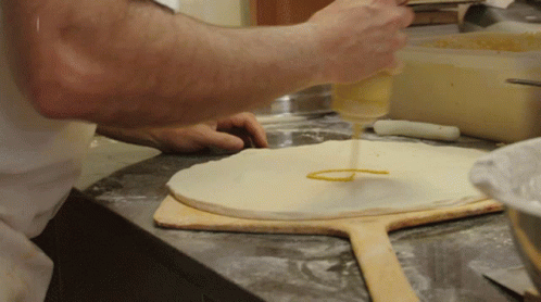 A Person Is Pouring Sauce On A Pizza Dough On A Wooden Paddle GIF