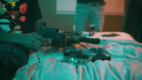 A Person Is Sitting On A Bed Holding A Gun And A Lighter . GIF