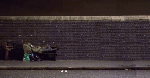A Person Is Sleeping On A Bench In Front Of A Brick Wall With A Trash Can On The Sidewalk GIF