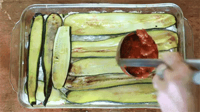 A Person Is Spreading Tomato Sauce On A Casserole Dish Filled With Zucchini Slices . GIF