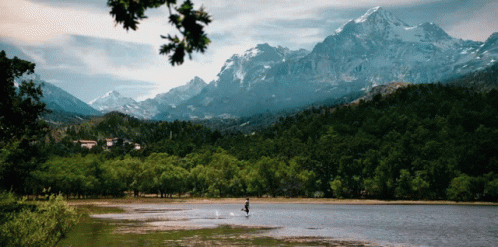 A Person Is Standing In The Middle Of A Lake With Snowy Mountains In The Background GIF