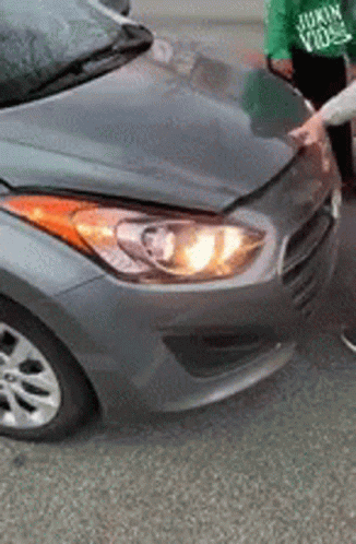 A Person Is Standing Next To A Gray Car With A Broken Hood And Headlight . GIF
