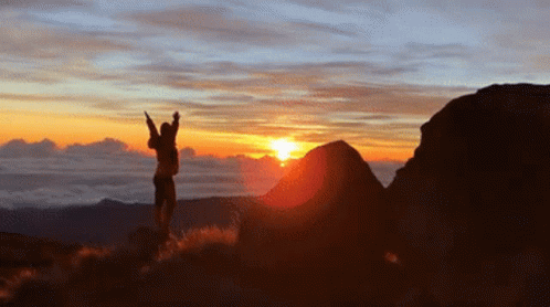 A Person Is Standing On Top Of A Hill With Their Arms In The Air At Sunset GIF