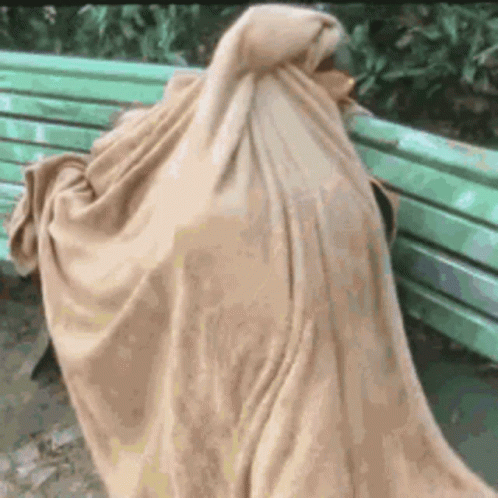 A Person Is Wrapped In A Blanket While Sitting On A Green Bench . GIF