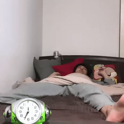 A Person Laying On A Bed Next To A Green Alarm Clock That Shows The Time As 7:15 GIF